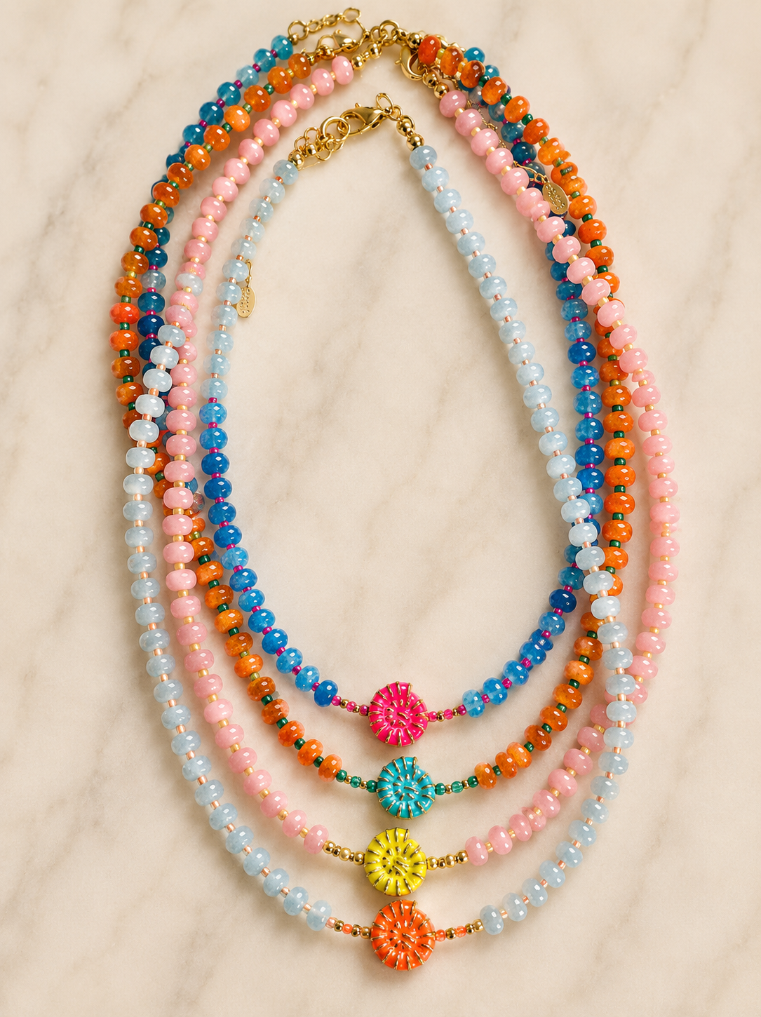 A set of five multi-strand beaded necklaces arranged in a fan shape on a light marble background. The necklaces feature pastel pink, light blue, and bright orange beads, with gold-tone clasp closures and decorative woven flower charms in pink, teal, yellow, and orange.