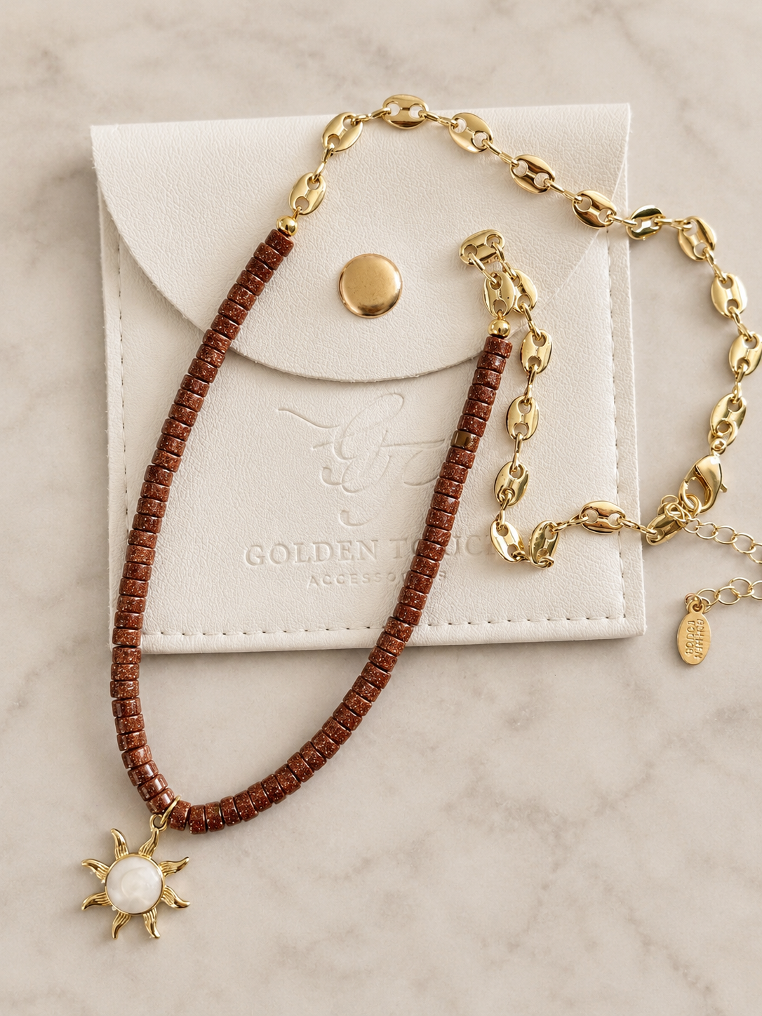 A gold chain necklace with a brown beaded strand and a sun pendant, displayed on a white leather jewelry pouch over a light marble surface.