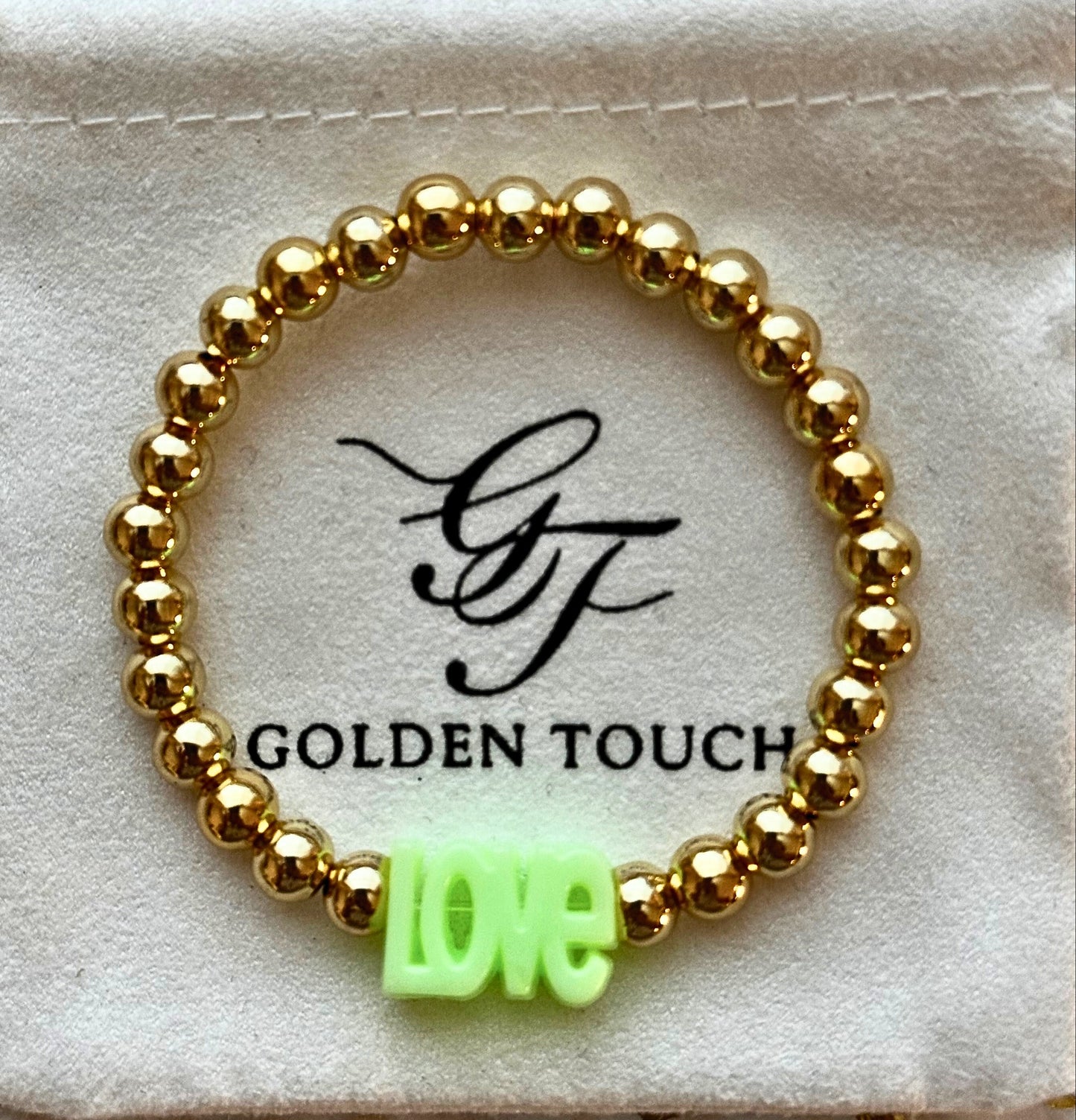 Gold beaded bracelet with a green 'LOVE' charm on a Golden Touch branded card.