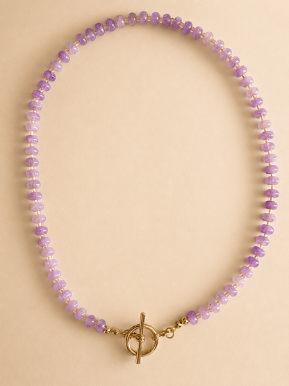 A beaded necklace made of small lavender and light purple faceted beads with a gold toggle clasp, displayed on a solid beige background.