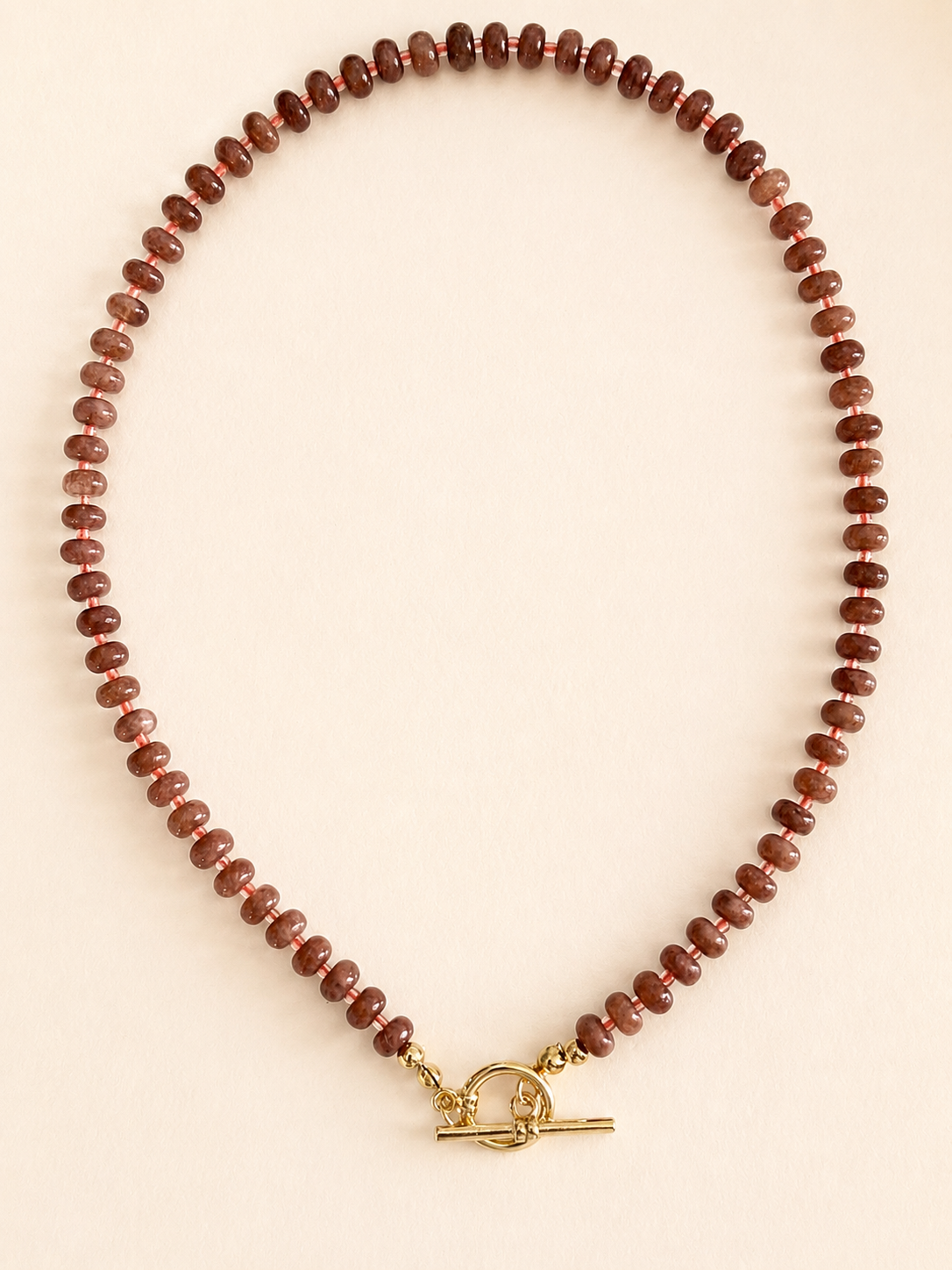 A beaded necklace made of small brown stone beads with a gold toggle clasp, displayed on a light beige background.