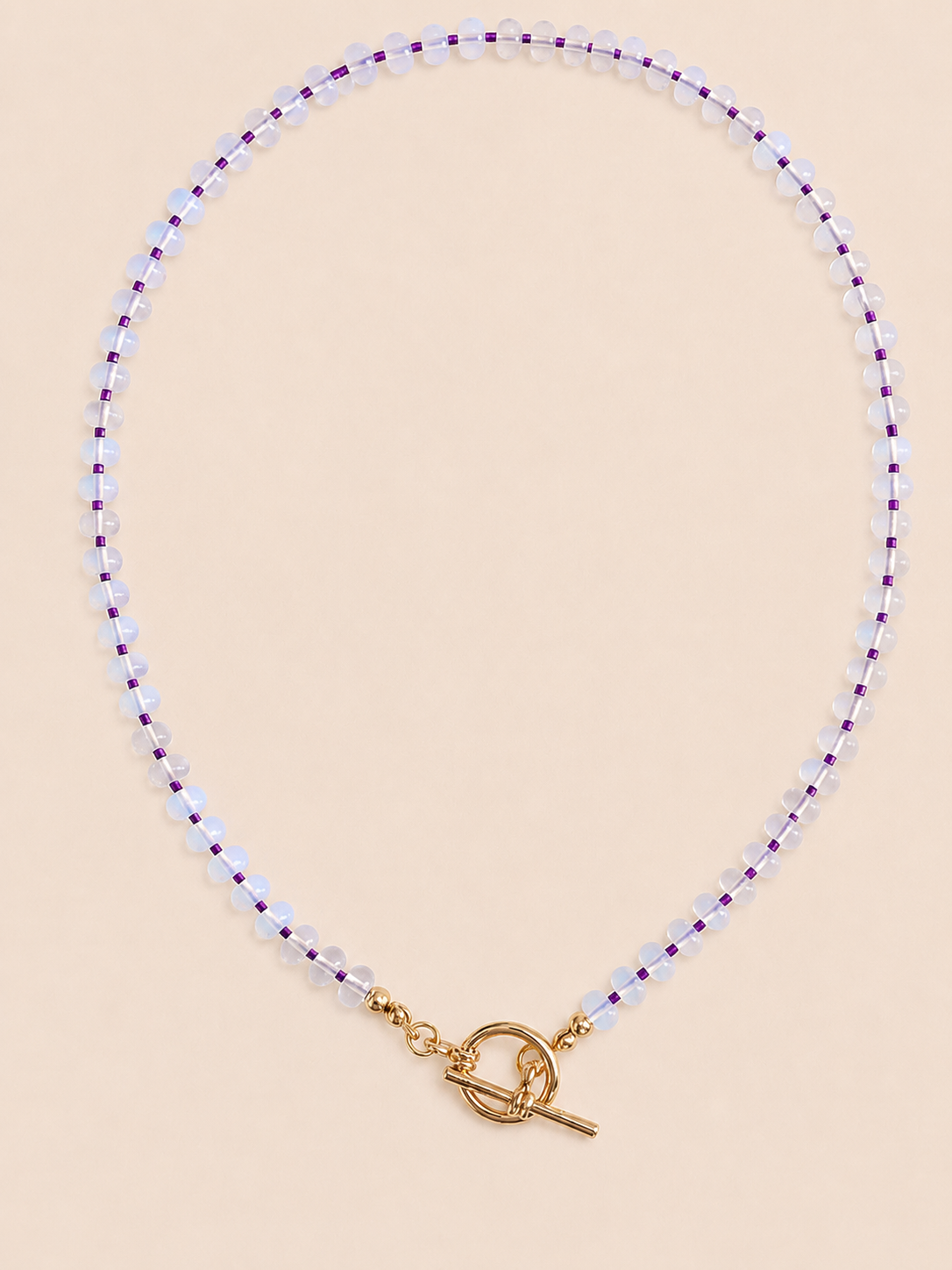 A delicate beaded necklace with small opal beads separated by thin purple seed beads, finished with a gold tbar pendant, displayed on a neutral beige surface.