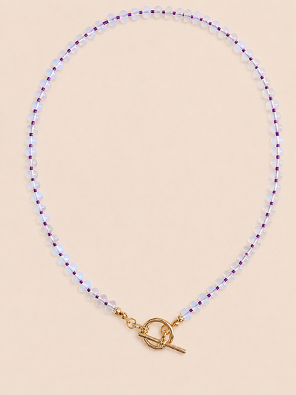 A delicate beaded necklace with small opal beads separated by thin purple seed beads, finished with a gold tbar pendant, displayed on a neutral beige surface.