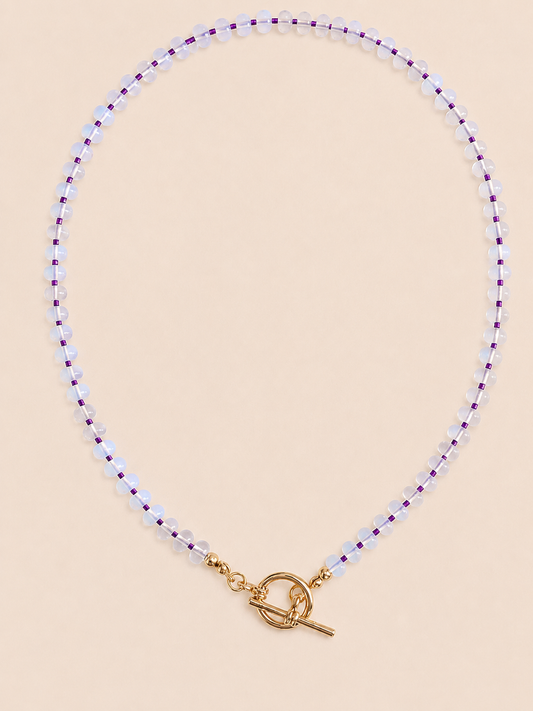 A delicate beaded necklace with small opal beads separated by thin purple seed beads, finished with a gold tbar pendant, displayed on a neutral beige surface.