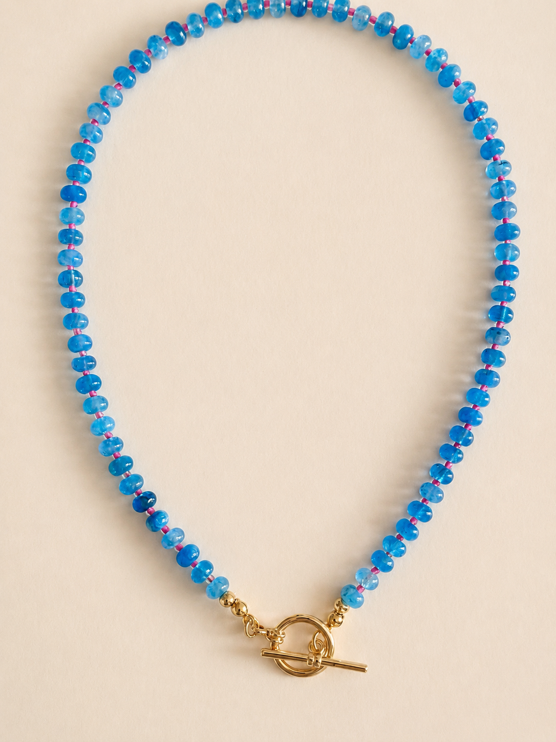 A beaded necklace made of small sky blue natural beads with a gold toggle clasp, photographed on a plain light beige background. Made by hand