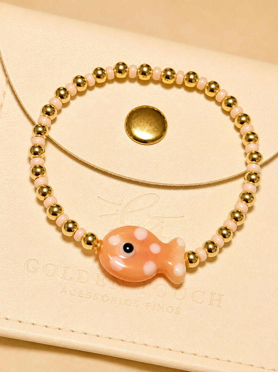 A delicate beaded bracelet with gold-toned round beads, pale pink translucent beads, and a central peach-colored fish charm with white spots, displayed on a cream-colored leather pouch.