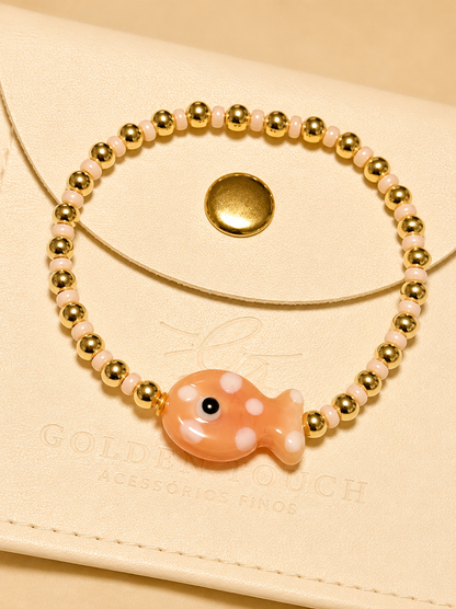 A delicate beaded bracelet with gold-toned round beads, pale pink translucent beads, and a central peach-colored fish charm with white spots, displayed on a cream-colored leather pouch.
