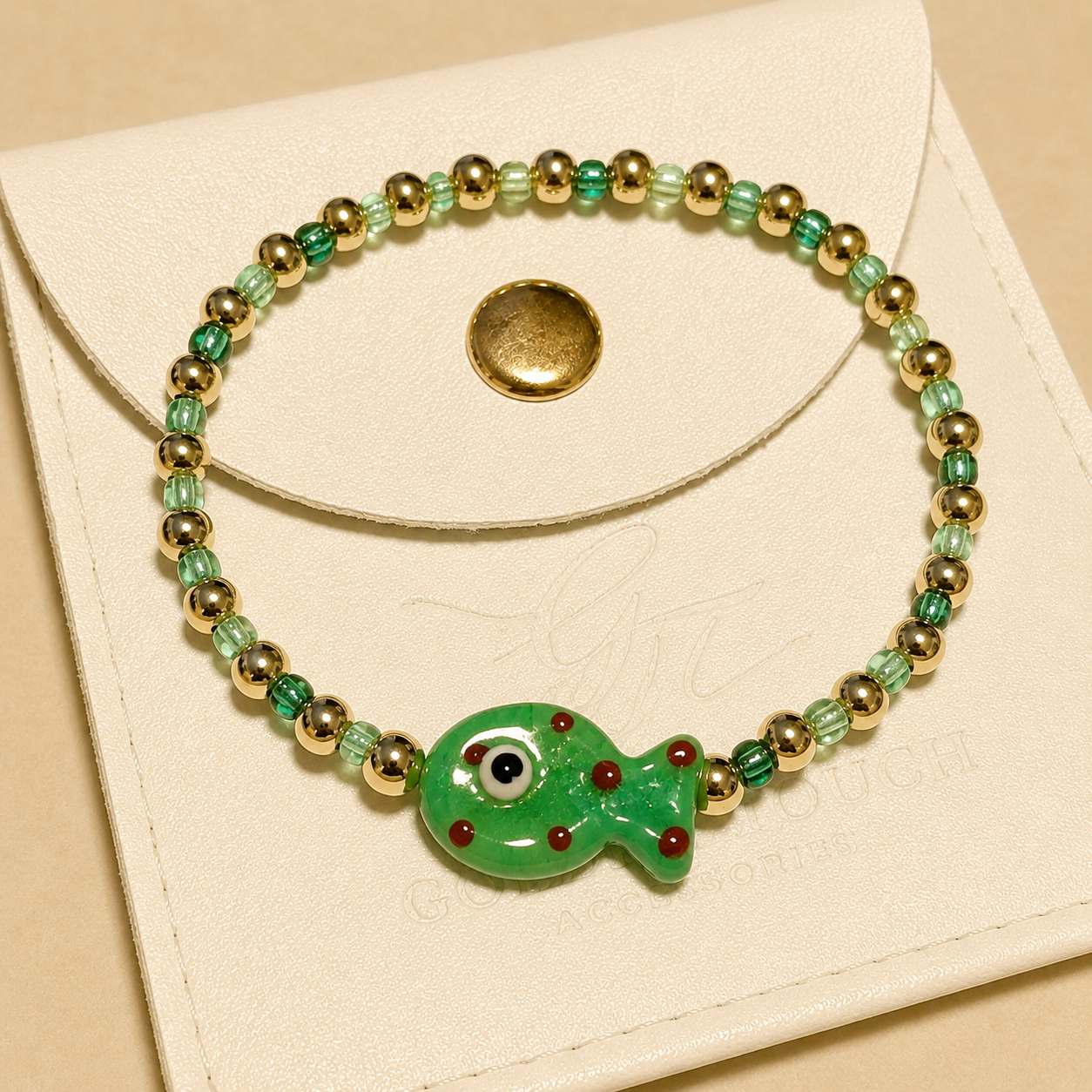 A beaded stretch bracelet with a green fish charm, displayed on a cream-colored leather pouch. The bracelet features green and light green beads, gold-toned spacer beads, and a central green fish charm with red details.