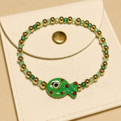 A beaded stretch bracelet with a green fish charm, displayed on a cream-colored leather pouch. The bracelet features green and light green beads, gold-toned spacer beads, and a central green fish charm with red details.