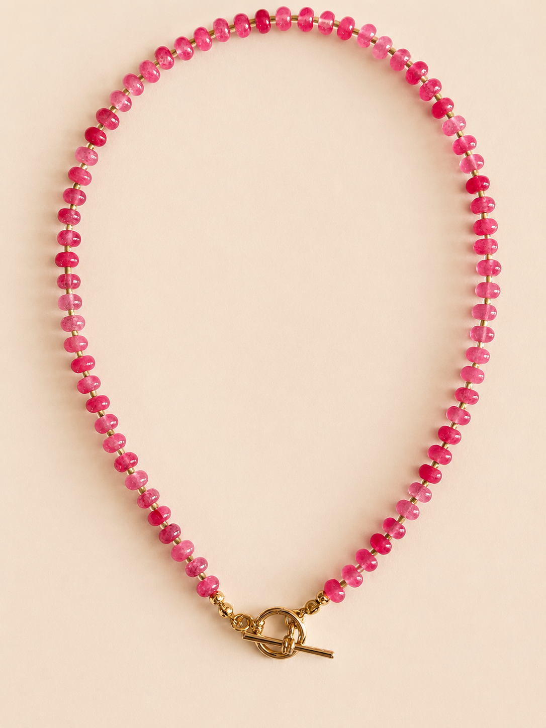 A beaded necklace made of small strawberry pink natural beads with a gold toggle clasp, displayed on a solid light beige background.