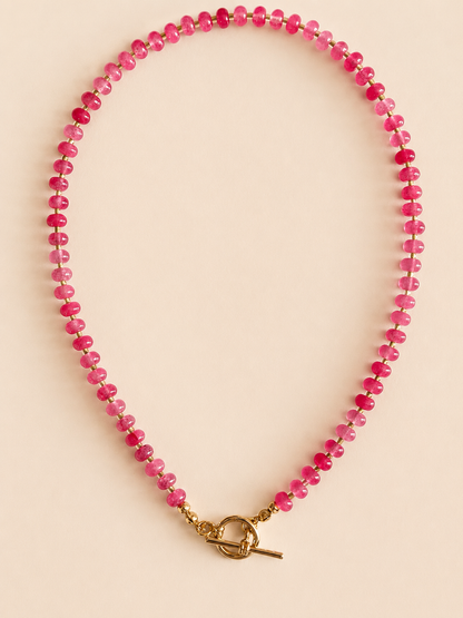 A beaded necklace made of small strawberry pink natural beads with a gold toggle clasp, displayed on a solid light beige background.