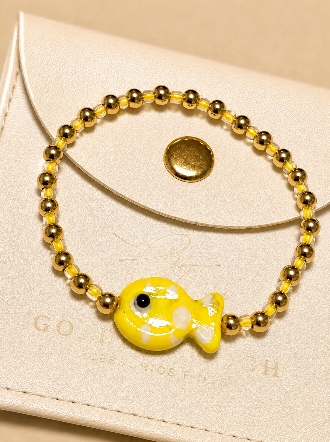 A gold beaded bracelet with a yellow and white fish charm, displayed on a cream-colored leather pouch.