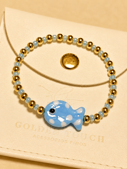 A beaded stretch bracelet with light blue beads, gold spacer beads, and a blue and white spotted fish charm, displayed on a cream-colored envelope with the brand name 'GOLDEN TOUCH' visible.