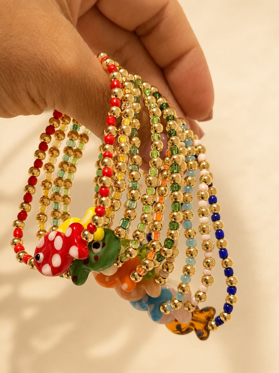 A hand holding a cluster of beaded necklaces with colorful frog and mushroom charms against a plain light background.