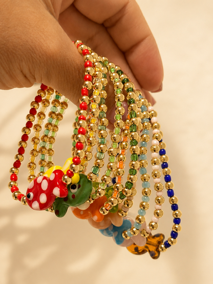 A hand holding a cluster of beaded necklaces with colorful frog and mushroom charms against a plain light background.