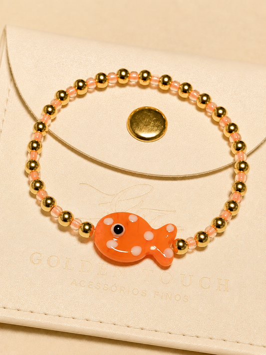 A beaded bracelet with gold-toned metal beads, neon orange translucent beads, and a glossy glass fish charm with white polka dots, displayed on a cream-colored envelope-style pouch.