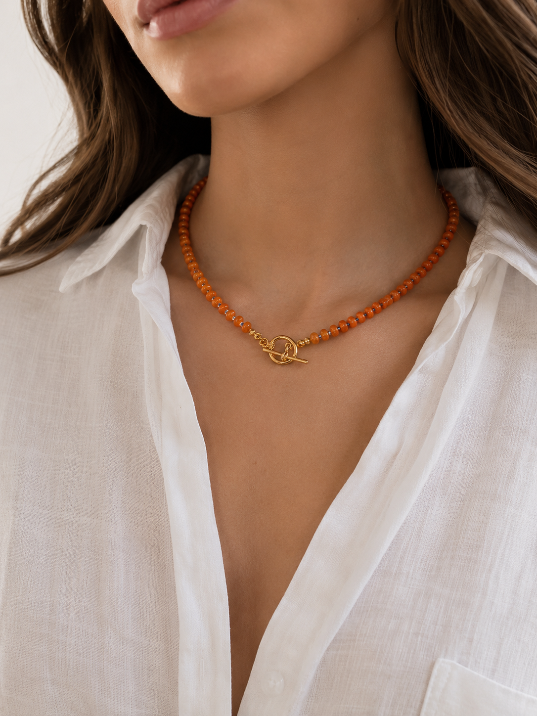 Close-up of a woman wearing a beaded orange necklace with a gold interlocking rings clasp, tbar styled with a white linen shirt.