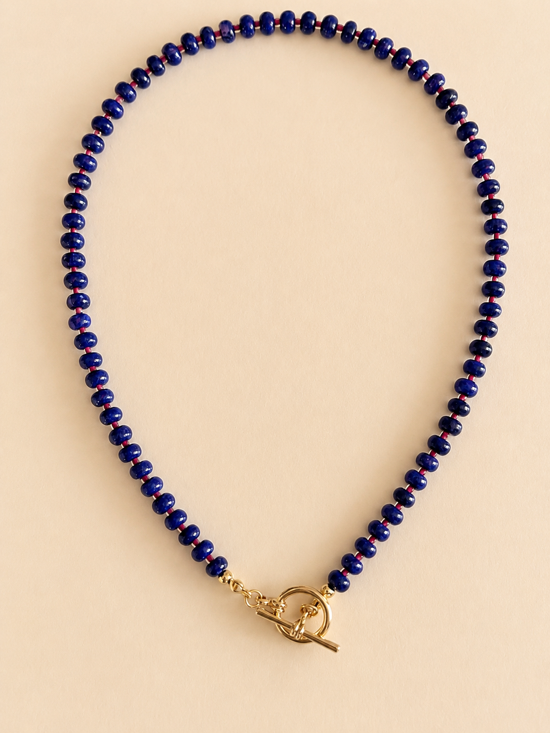 A beaded necklace made of small dark blue beads with a gold toggle clasp, displayed on a solid light beige background. tbar design