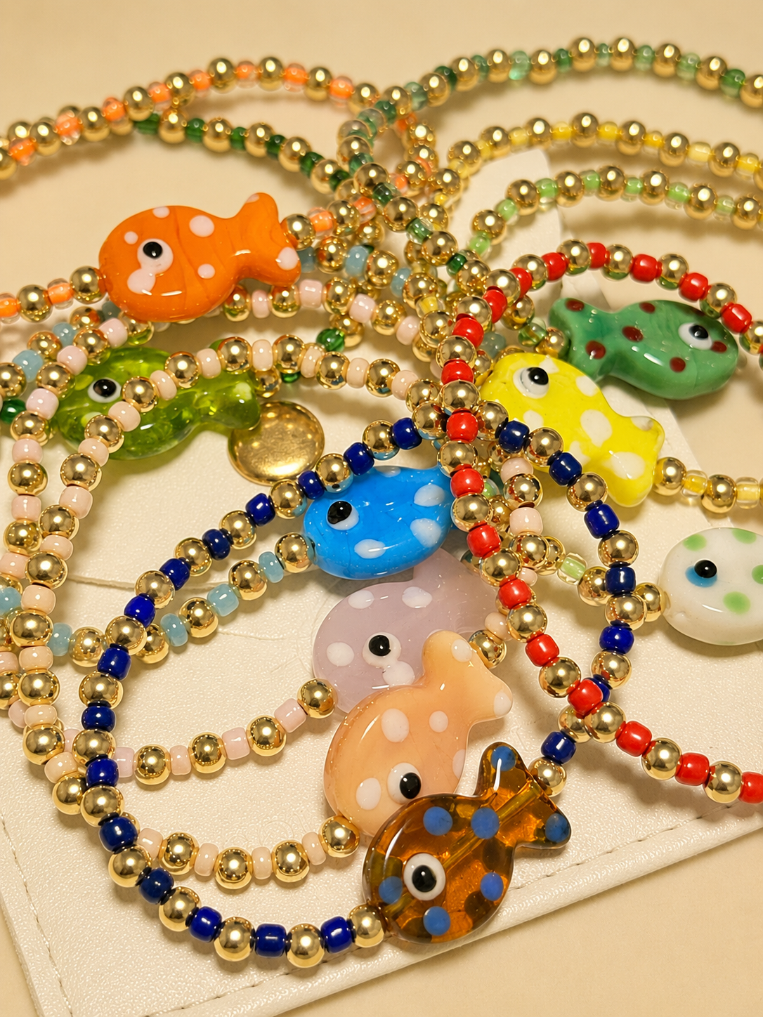 A close-up of several beaded necklaces or bracelets arranged on a light beige surface. The strands are made of small metallic gold beads with colorful round beads in blue, red, green, orange, yellow, and white, and they feature glossy fish-shaped charms in various colors.