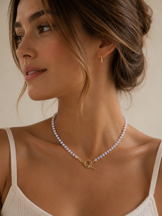 A close-up of a woman wearing a delicate opal necklace with a tbar front closure and a small gold hoop earring, set against a warm beige background.