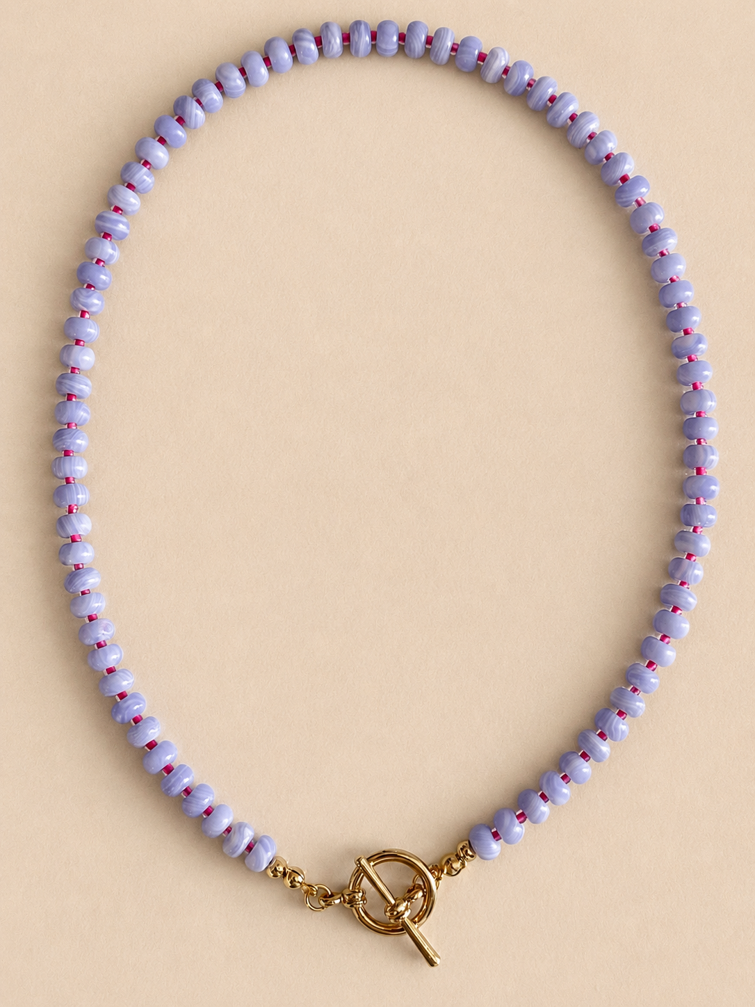 A beaded necklace with small lavender and white beads, each detailed with tiny red dots, featuring a gold toggle clasp, displayed on a solid beige background.