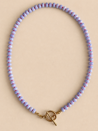 A beaded necklace with small lavender and white beads, each detailed with tiny red dots, featuring a gold toggle clasp, displayed on a solid beige background.