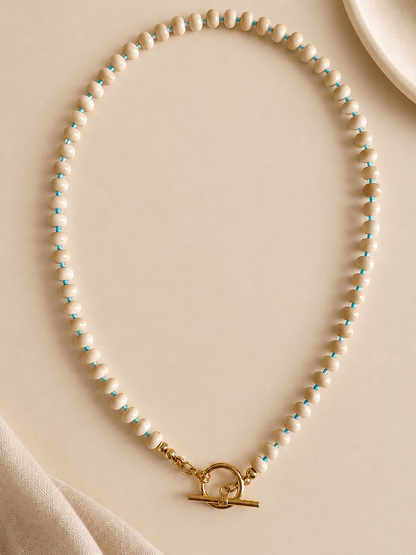 A delicate beaded necklace with small cream agate beads separated by thin turquoise blue seed beads, finished with a gold tbar pendant, displayed on a neutral beige surface.