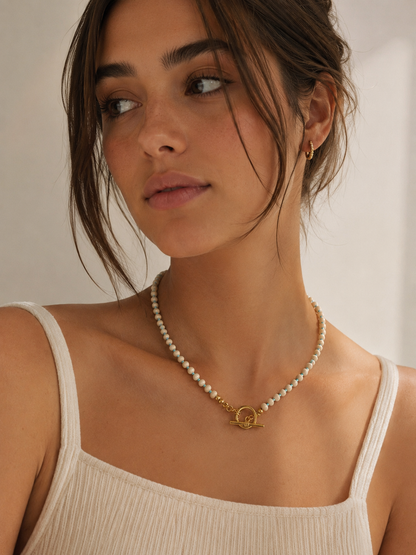 A close-up of a woman wearing a delicate cream agate beaded necklace with a gold-tone circular interlocking rings clasp, paired with a cream ribbed tank top.