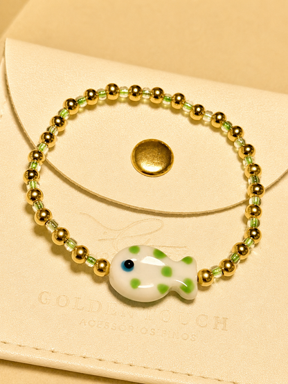 A beaded stretch bracelet with gold-toned round beads, light green faceted beads, and a white fish charm with green spots and a blue eye, displayed on a cream-colored leather pouch.