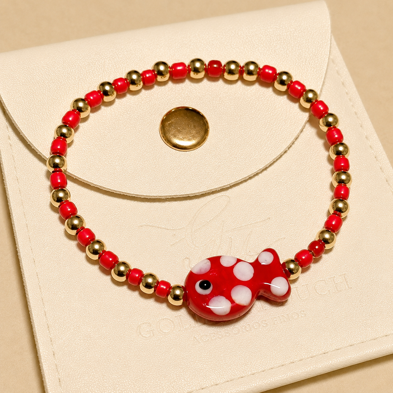 A beaded stretch bracelet with red and white beads, gold spacer beads, and a red fish charm with white polka dots, displayed on a cream-colored leather pouch.