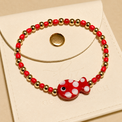 A beaded stretch bracelet with red and white beads, gold spacer beads, and a red fish charm with white polka dots, displayed on a cream-colored leather pouch.