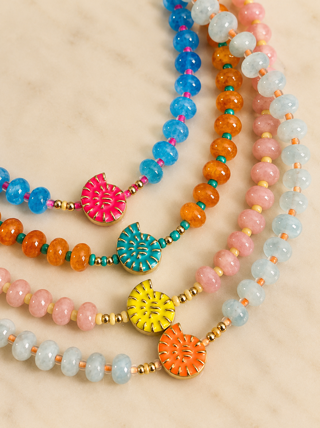 Four beaded necklaces arranged in a fan shape on a light beige marble surface. The necklaces feature faceted round beads in blue, orange, pink, and light aqua, separated by small gold spacer beads, and decorated with colorful enamel lotus-shaped charms in pink, teal, yellow, and orange.