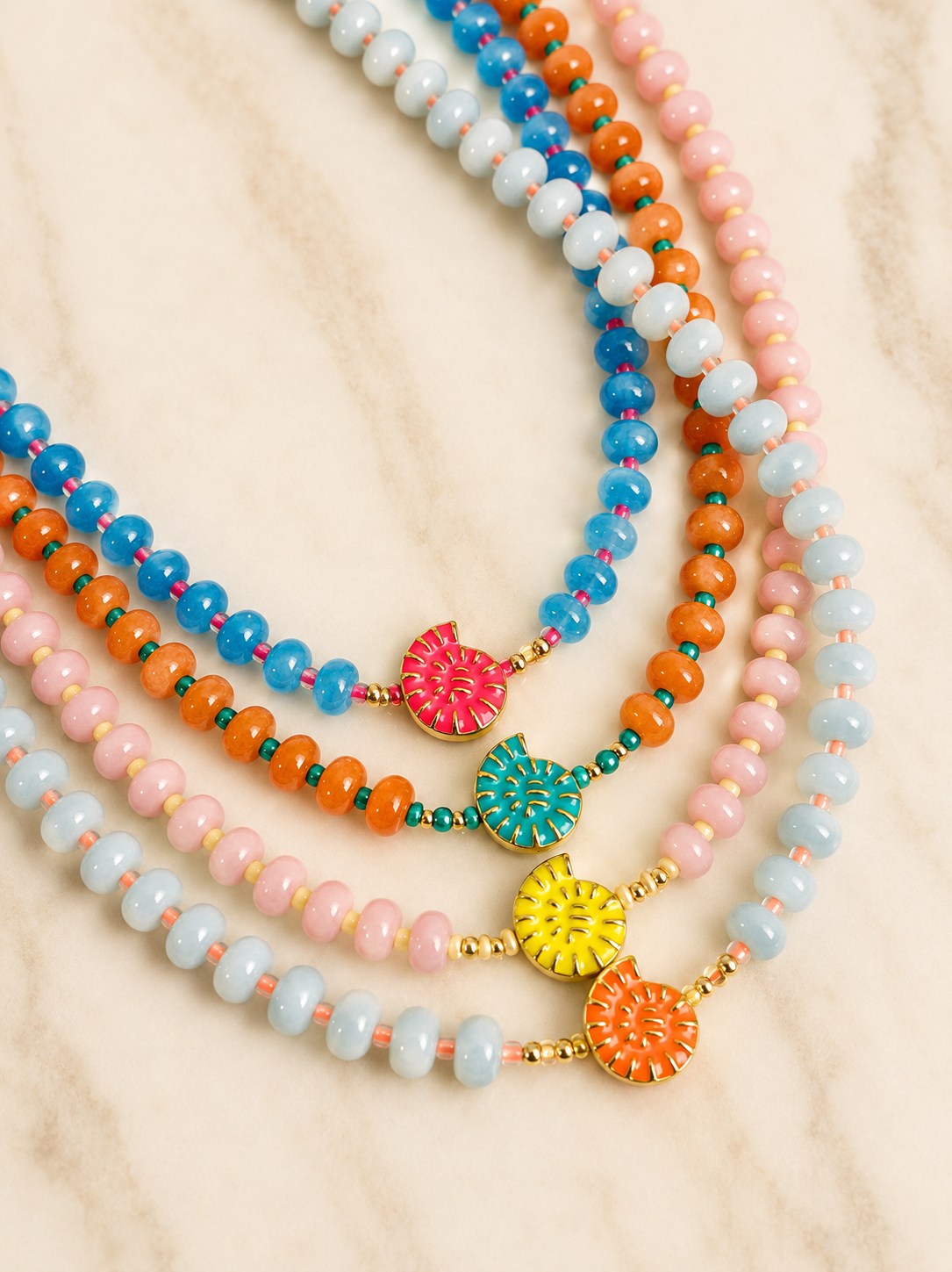 Four beaded necklaces in pastel blue, orange, pink, and light aqua, each with a colorful flower-shaped charm and gold-tone accents, arranged on a light marble surface.