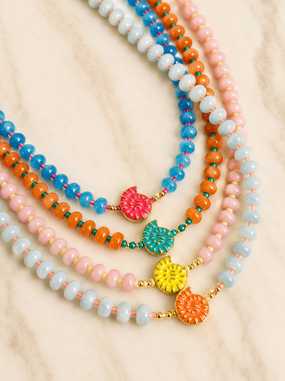 Four beaded necklaces in pastel blue, orange, pink, and light aqua, each with a colorful flower-shaped charm and gold-tone accents, arranged on a light marble surface.