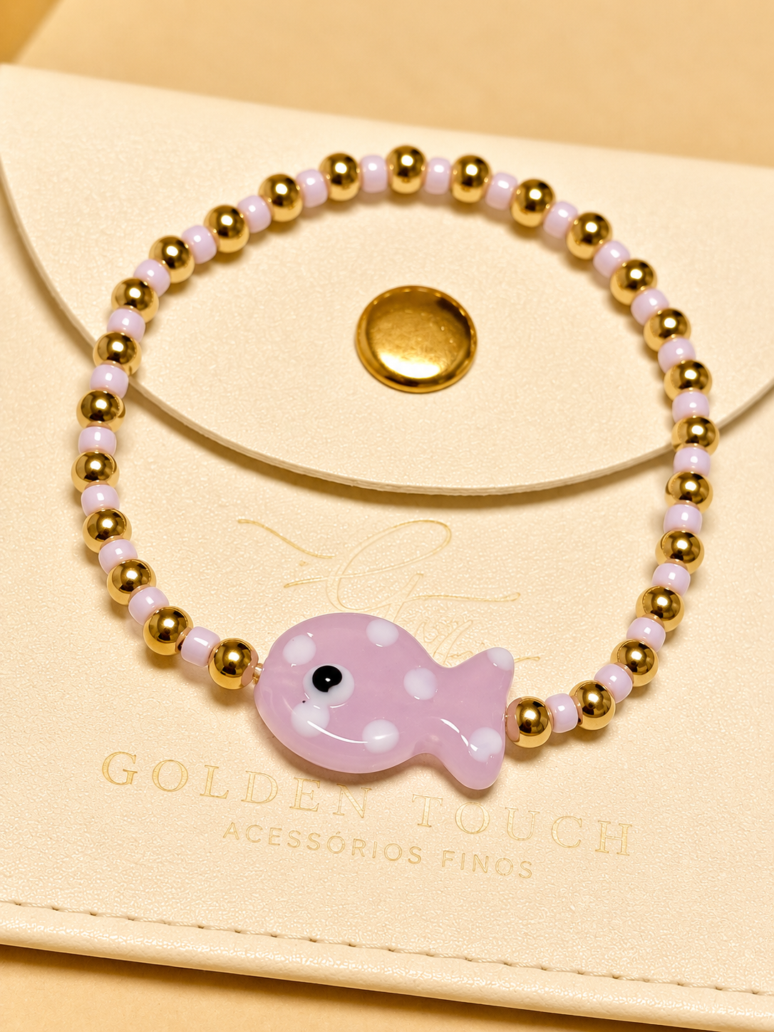 A beaded stretch bracelet with gold-toned round beads, pale pink translucent beads, and a central light pink fish charm with white polka dots and a black eye, displayed on a cream-colored card with the brand name Golden Touch.
