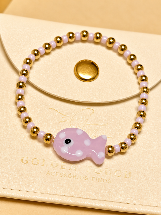 A beaded stretch bracelet with gold-toned round beads, pale pink translucent beads, and a central light pink fish charm with white polka dots and a black eye, displayed on a cream-colored card with the brand name Golden Touch.