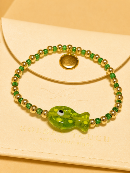 A green beaded bracelet with a translucent green fish charm and gold-tone spacer beads, displayed on a cream-colored envelope with the brand name 'Goldie Touch' partially visible.