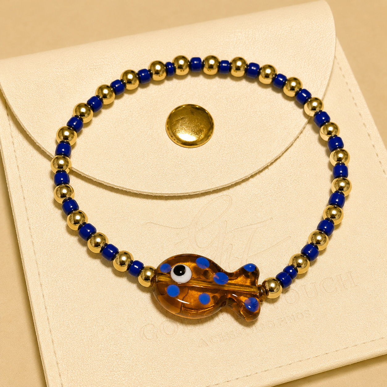 A beaded stretch bracelet with alternating royal blue and metallic gold beads, featuring a translucent amber-brown evil eye fish charm in the center, displayed on a cream-colored envelope-style card.
