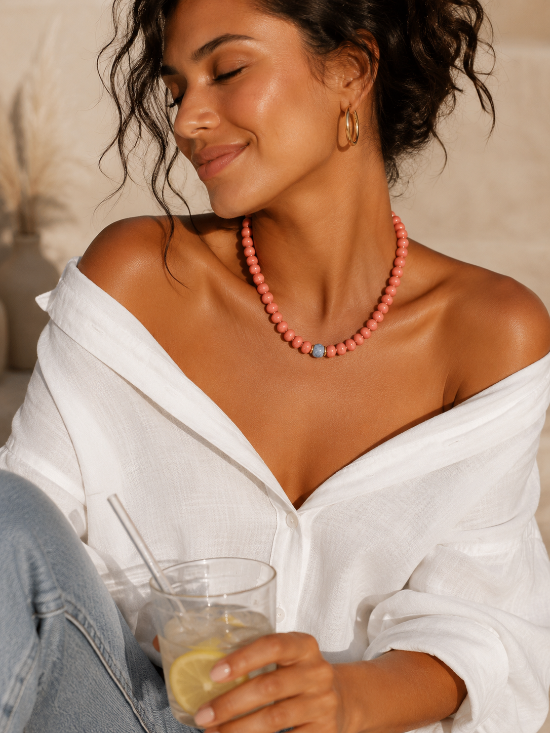 Woman wearing a burnt orange beaded necklace, holding a drink, with a neutral background