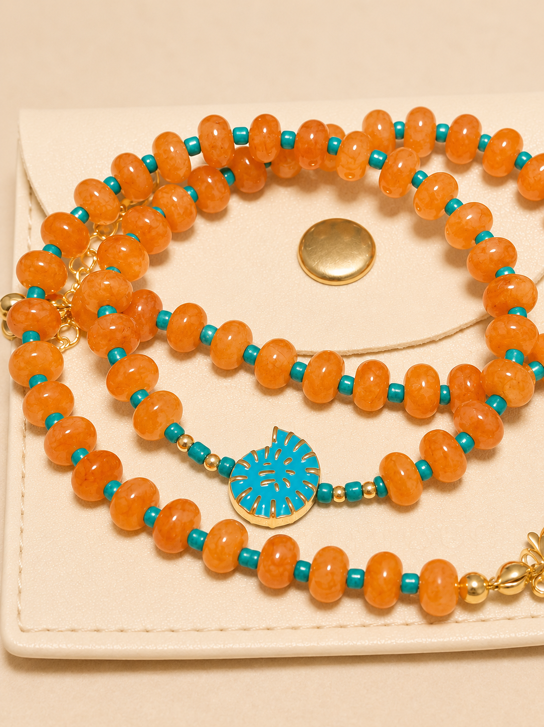 A beaded necklace with orange marbled beads, turquoise blue spacer beads, and a central turquoise and gold sunburst medallion, displayed on a cream-colored textured card.