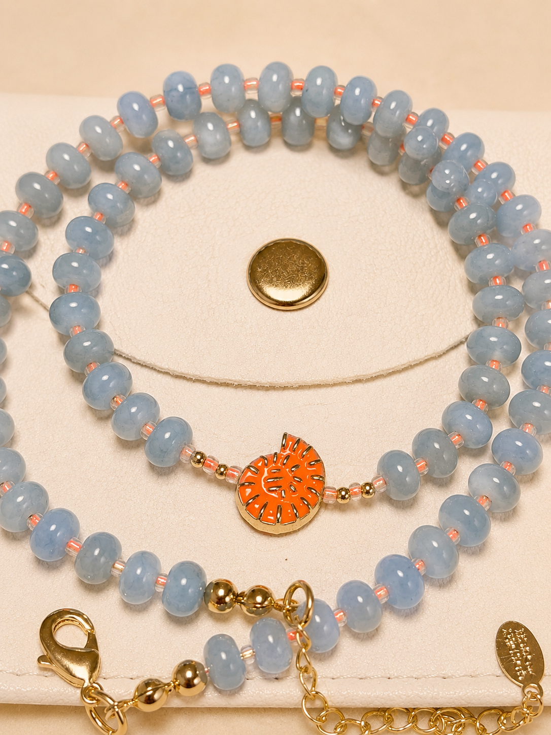 A beaded necklace set displayed on a cream jewelry stand. The set features light blue faceted beads separated by small peach-pink spacer beads, with a central orange and gold patterned heart charm.