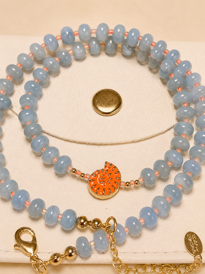 A beaded necklace set displayed on a cream jewelry stand. The set features light blue faceted beads separated by small peach-pink spacer beads, with a central orange and gold patterned heart charm.