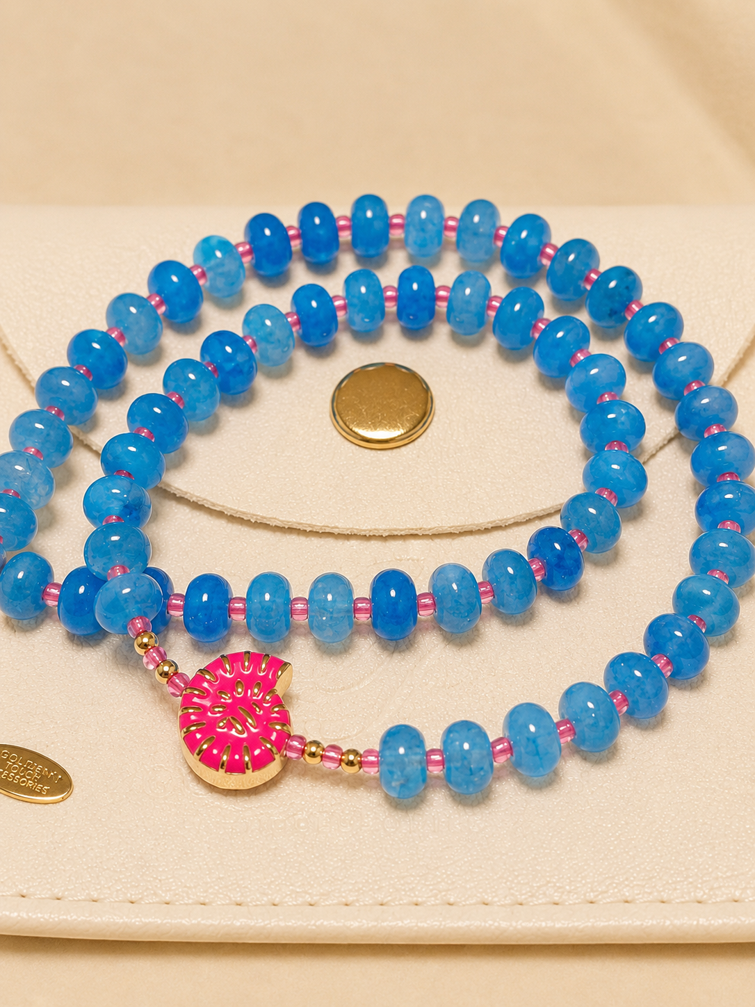 A beaded necklace with round blue marbled beads separated by thin pink threads, featuring a gold-tone circular pendant with pink floral detailing, displayed on a cream-colored leather pouch.