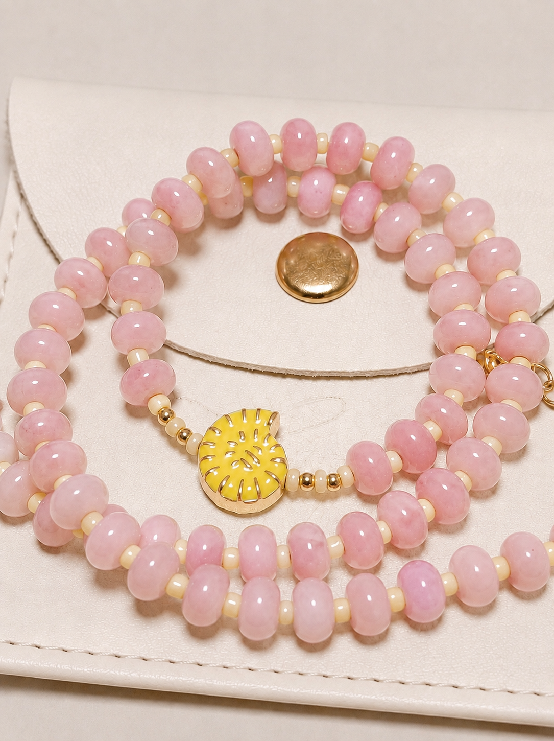 A close-up of a multi-strand beaded necklace featuring light pink stone beads, small cream spacer beads, and a textured yellow crescent moon charm with gold detailing, displayed on a white leather surface.