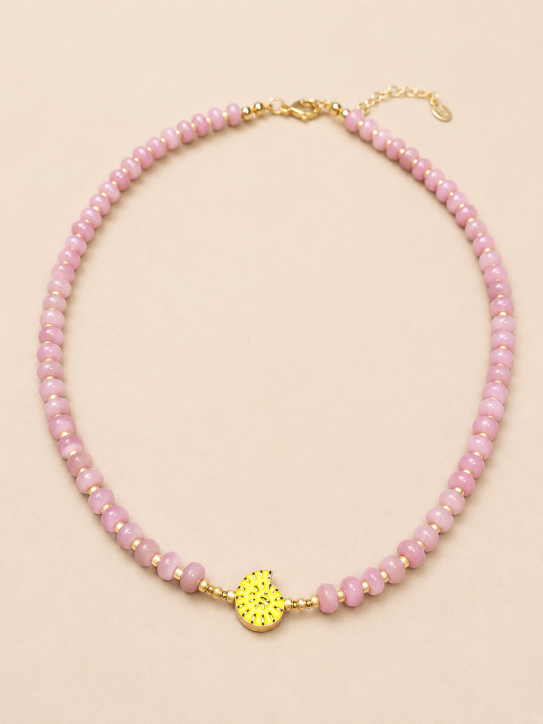 A dusty pink beaded necklace with a textured yellow gold shell charm in the center, displayed on a solid light beige background.