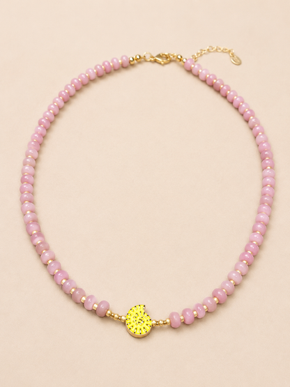 A dusty pink beaded necklace with a textured yellow gold shell charm in the center, displayed on a solid light beige background.
