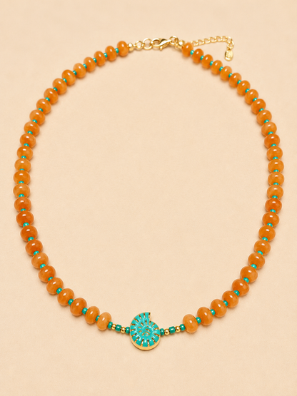 A beaded necklace featuring round orange agate beads accented with small turquoise blue beads, a central turquoise shell shaped charm with gold detailing, and a gold clasp on a light beige background.