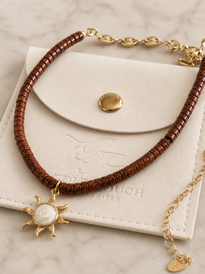 A brown beaded necklace with a gold sun pendant featuring a white pearl center, displayed on a cream-colored leather pouch over a light marble surface.