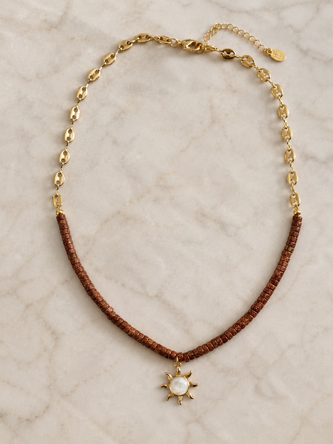 A gold chain necklace with sunstone brown beaded section and a small gold sun pendant featuring a white pearl center, displayed on a light marble surface.