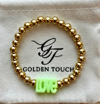Gold beaded bracelet with a green 'LOVE' charm on a Golden Touch branded card.